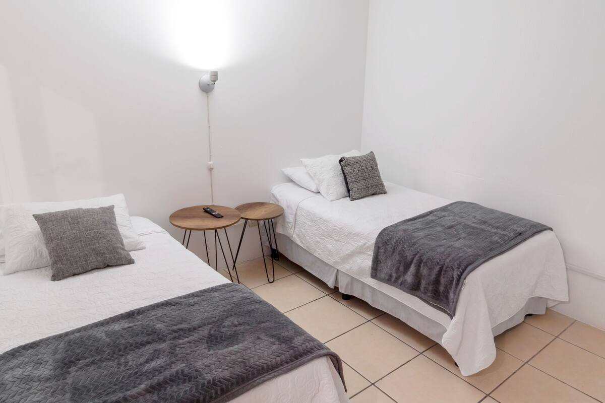 A room with two beds, a small table, and a wall-mounted light.