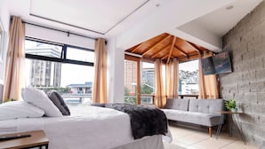 A modern hotel room with a large bed, a sofa, a TV mounted on a brick wall, and a view of the city through large windows. - Hotel Citadin Z4 (Guatemala City)