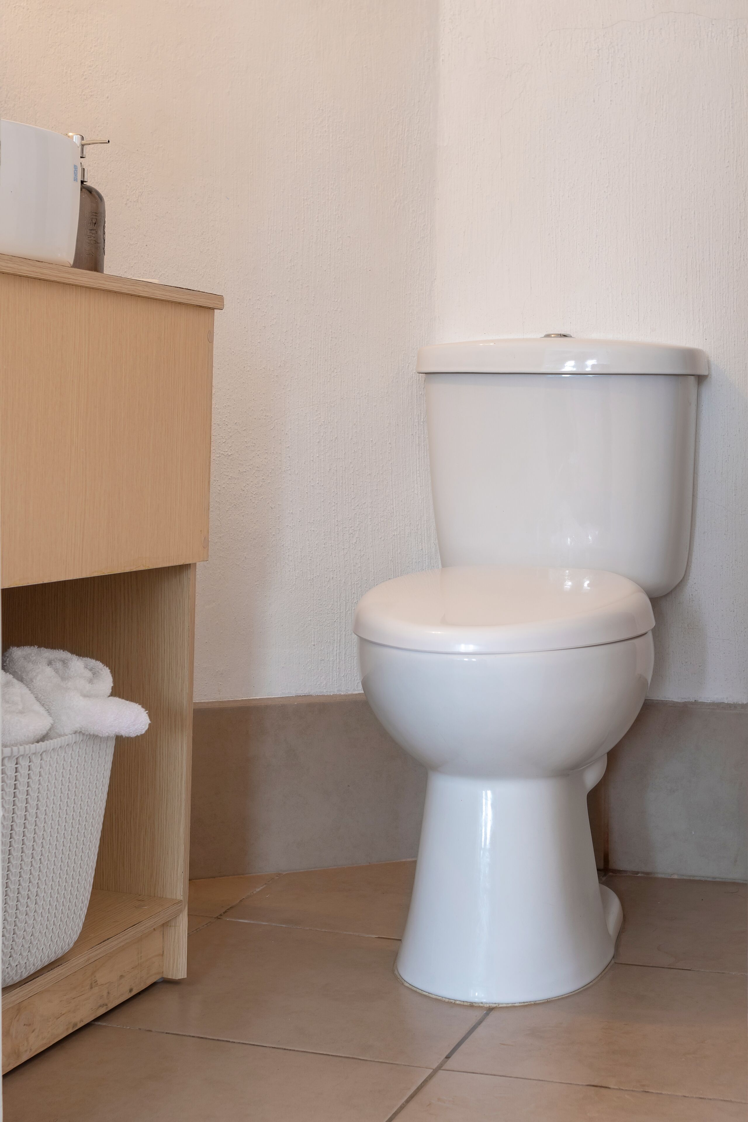 A white toilet with a tank, a wooden shelf with a white basket, and a beige tiled floor.