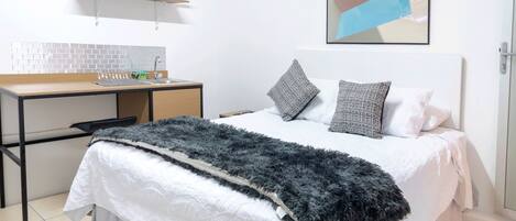 Un dormitorio con una cama, un escritorio, una silla, un estante con utensilios de cocina y un cuadro en la pared.