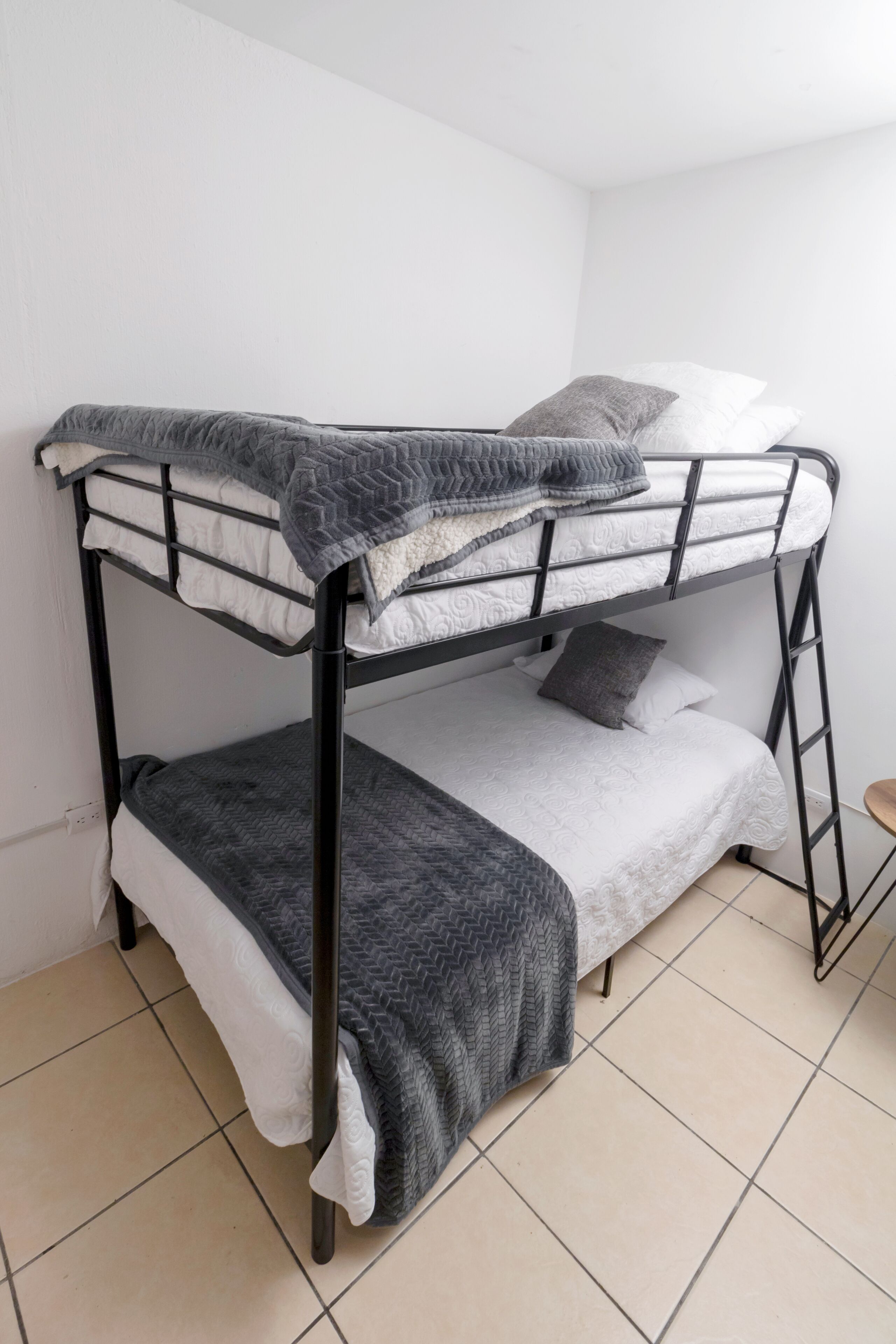 A bunk bed with a gray quilt and pillows, a small wooden stool, and a tiled floor.