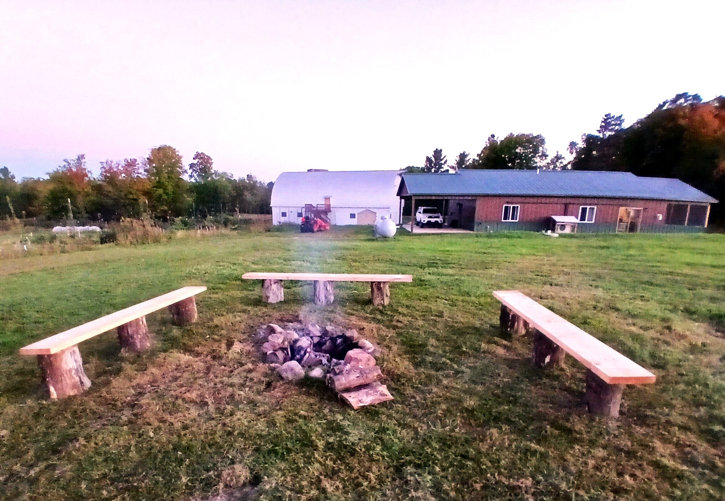 En gräsklädd äng med picknickbord och en eldstad, omgiven av lador och träd.