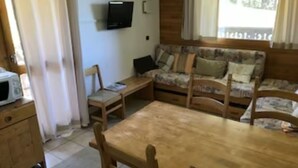 A cozy living room with a wooden table and chairs, a sofa with cushions, a television on the wall, and a window with curtains. - 2 bed Apt in Courchevel 1650, Doorstep skiing - Courchevel, Meribel, Val Thorens (Courchevel)