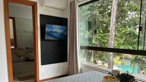 A bedroom with a bed, a TV mounted on the wall, a window with a view of trees and a pool, and a bedside table with a bowl of fruit and a plant.