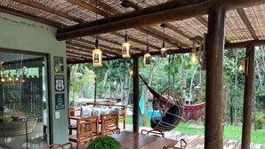 An outdoor dining area with a wooden table and chairs, a hanging chair, and a view of the surrounding greenery.
