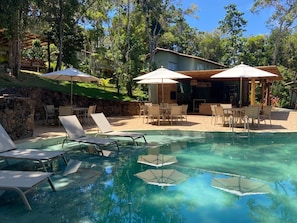 A clear swimming pool with lounge chairs and umbrellas, surrounded by a stone wall and trees. - House in Praia do Forte with a private lagoon. (Açu da Torre)