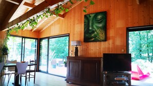 A spacious room with wooden walls and ceiling, a large window, a television, and a cabinet. - Pleasant wooden house near beaches and forest (Le Porge)