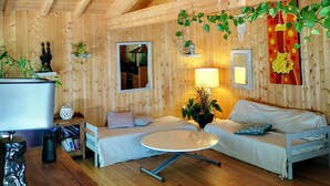 A cozy wooden cabin interior with a wooden ceiling, two sofas, a round table, and various decorations including plants and artwork. - Pleasant wooden house near beaches and forest (Le Porge)