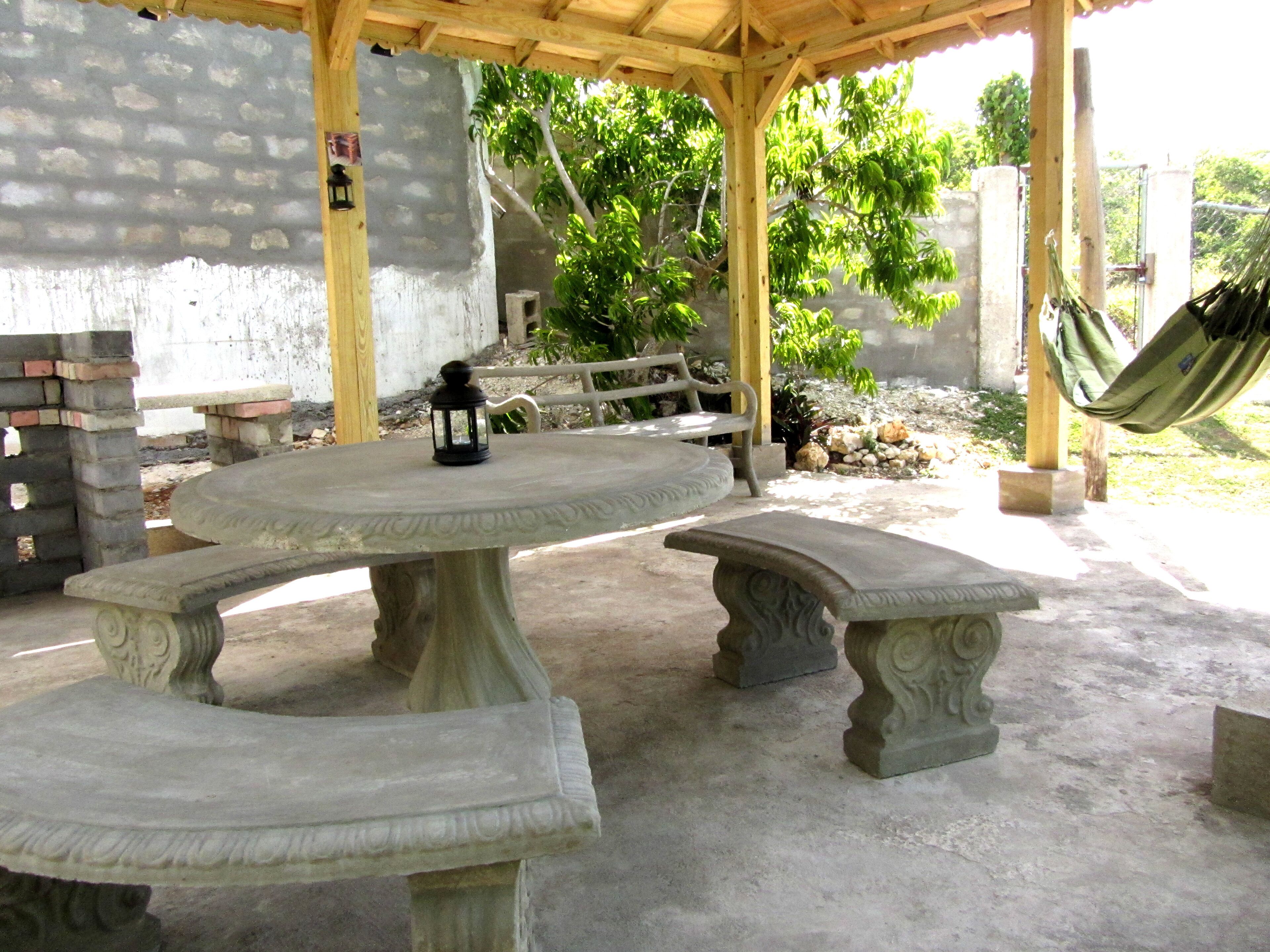 Un patio techado al aire libre con una mesa y bancos de piedra, rodeado por un muro de piedra y vegetación.