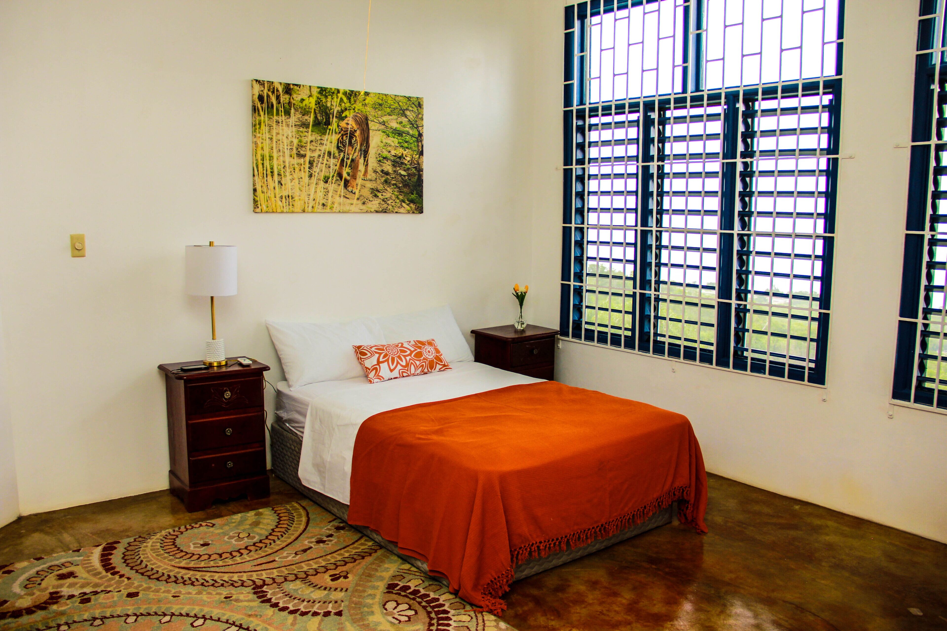 Un dormitorio con cama, mesita de noche, lámpara, ventana con rejas y un cuadro en la pared.