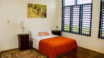 Un dormitorio con cama, mesita de noche, lámpara, ventana con rejas y un cuadro en la pared.
