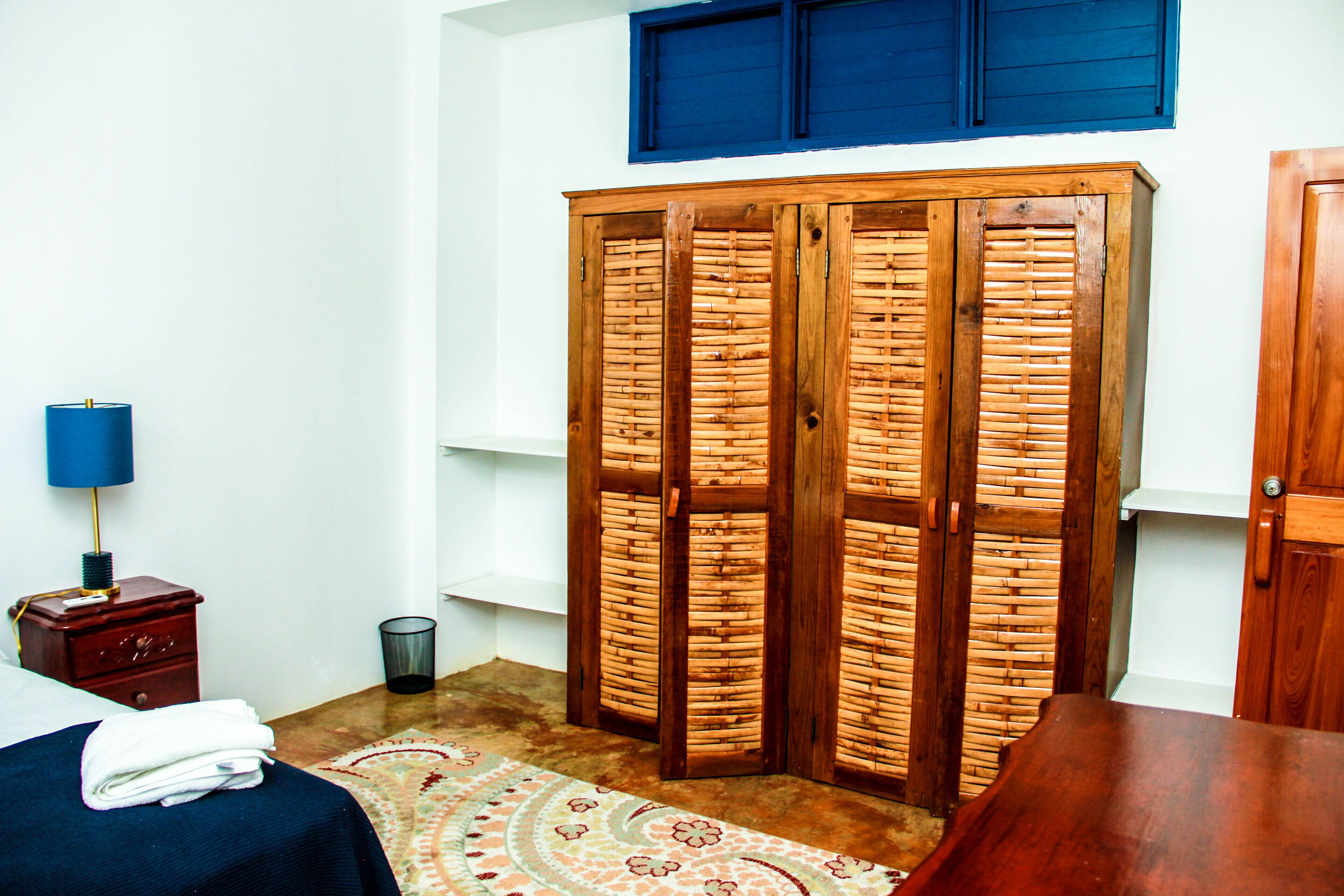 Una habitación con cama, armario, una mesita con lámpara y una alfombra estampada.