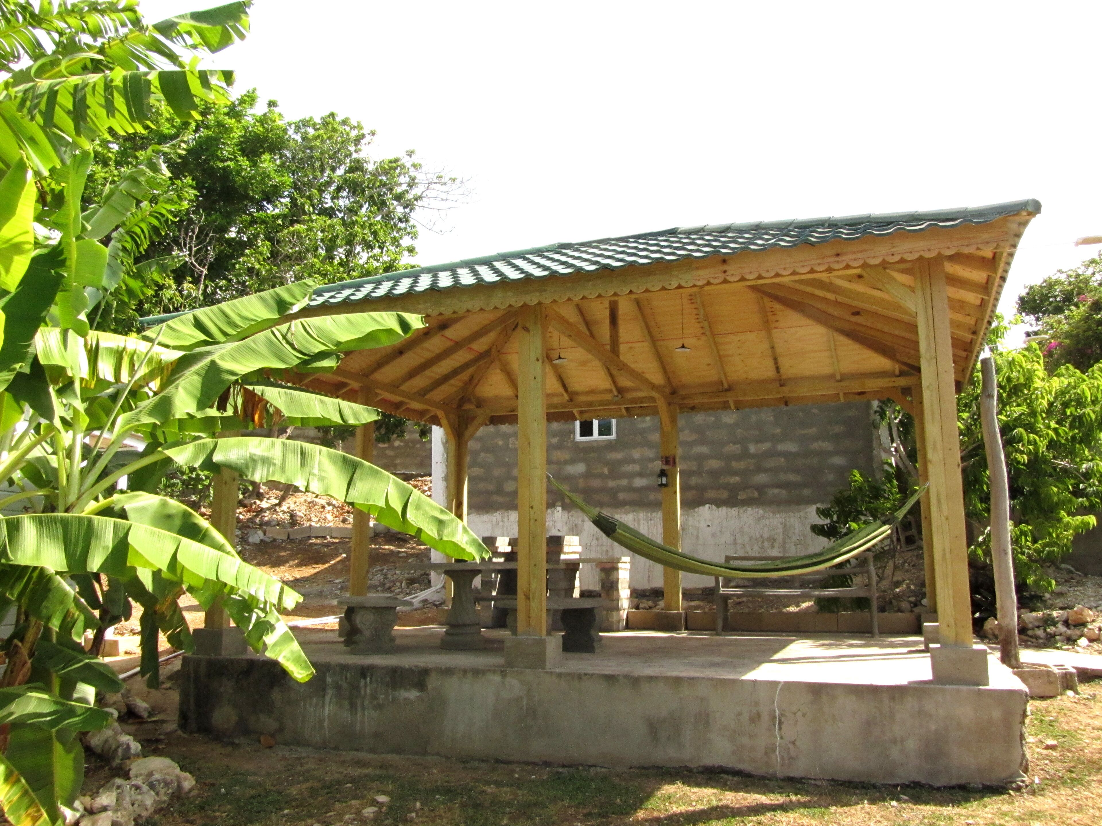 A wooden pavilion with a tiled roof, surrounded by greenery and a hammock.