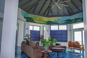 Une pièce dotée d’un ventilateur de plafond, d’un canapé, d’une table basse et d’un fauteuil. Les fenêtres sont ornées de barreaux bleus et le plafond est décoré d’une fresque représentant un paysage.