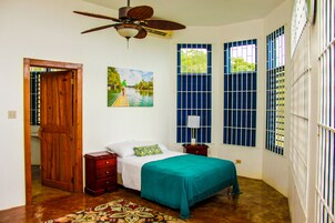 Un dormitorio con una cama, una puerta de madera, un ventilador de techo, una ventana con rejas y un cuadro en la pared.