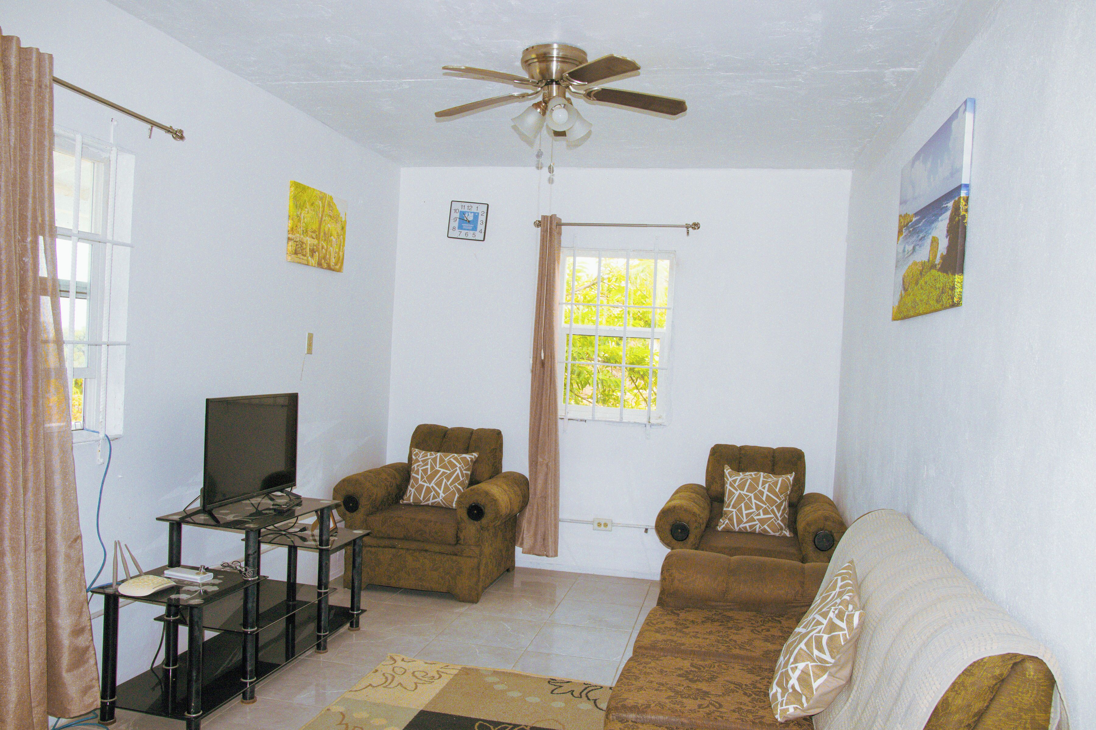 A living room with a ceiling fan, two armchairs, a sofa, a television on a stand, and a window with curtains.