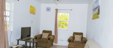 A living room with a ceiling fan, two armchairs, a sofa, a television on a stand, and a window with curtains.