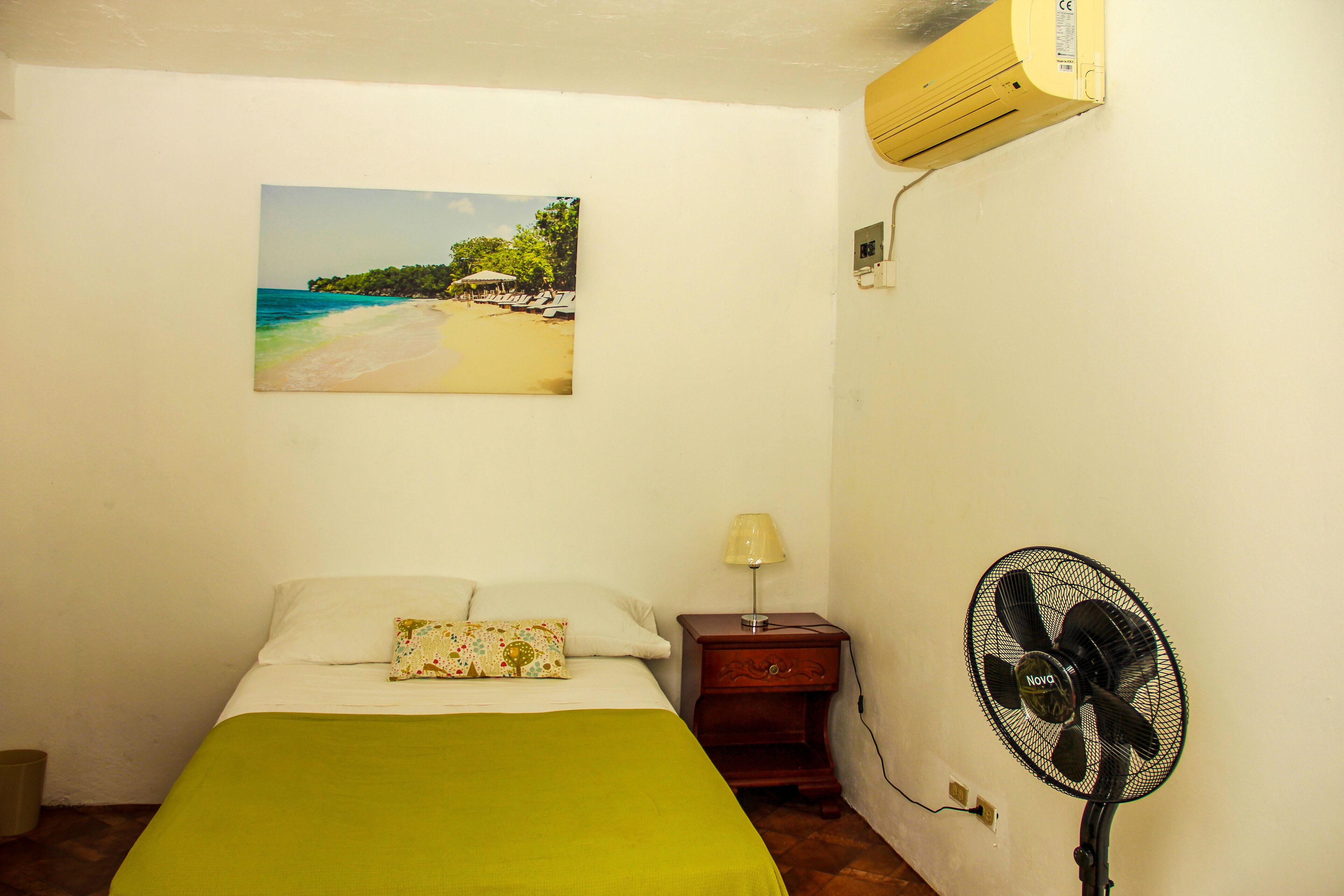 A bedroom with a bed, a nightstand, a wall-mounted air conditioner, a wall-mounted fan, and a beach scene painting.