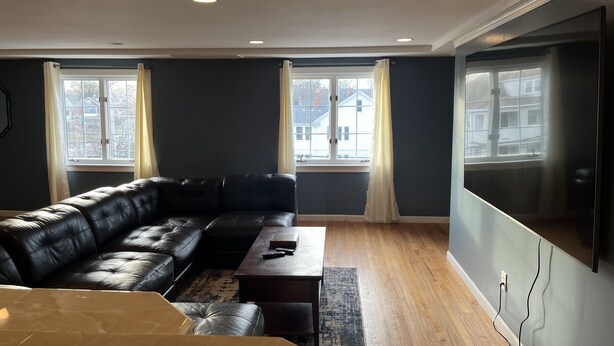 Una sala de estar con un sofá de cuero negro, una mesa de centro de madera y un televisor de pantalla plana grande montado en la pared. Hay dos ventanas con cortinas y el suelo es de madera.