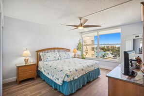 Quarto com cama, mesa de cabeceira, luminária, ventilador de teto, televisão e janela com vista para edifícios.