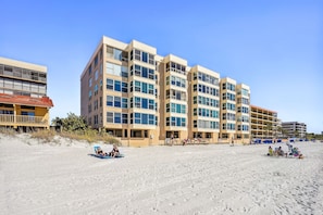A beachfront hotel with multiple floors, balconies, and a sandy beach area. - Madeira Vista 601-Corner Unit, Panoramic View (Madeira Beach)