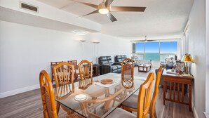 A spacious living room with a glass dining table, wooden chairs, a ceiling fan, and a view of the ocean. - Madeira Vista 601-Corner Unit, Panoramic View (Madeira Beach)