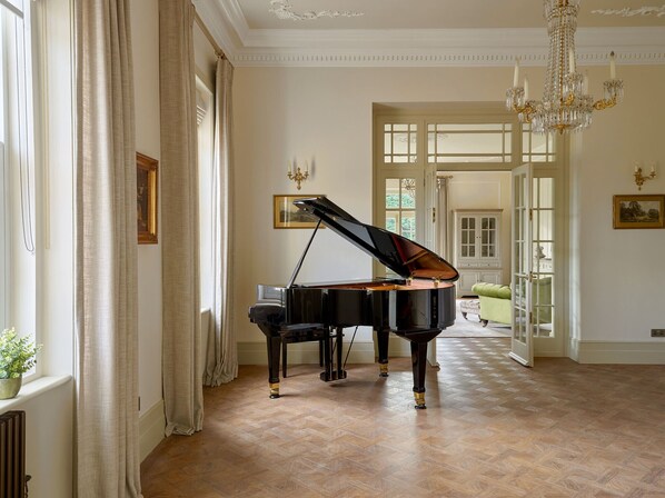Un piano de cola en una habitación amplia con piso de parquet, ventanas grandes con cortinas y un candelabro.