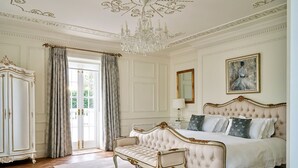 Ein luxuriöses Schlafzimmer mit einem großen Bett, einer Liege, einem klassischen Kronleuchter und Blick ins Freie.