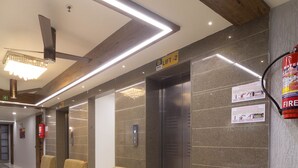 A hotel lobby with a reception desk, a fire extinguisher, a sign indicating a lift, and a fire escape door. - Hotel Green City (Gandhinagar)