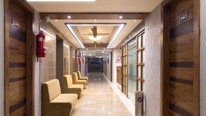 A hotel corridor with a row of beige sofas, wooden paneling on the walls, and a shiny tiled floor. - Hotel Green City (Gandhinagar)