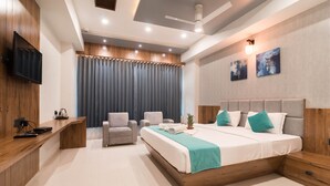 A modern hotel room with a wooden headboard, a large bed, a desk with a flat-screen TV, and a seating area with two armchairs. - Hotel Green City (Gandhinagar)