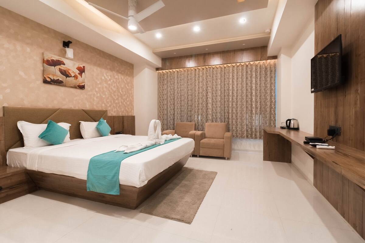 A modern hotel room with a large bed, a seating area with a chair, a desk with a coffee maker, and a TV mounted on the wall.
