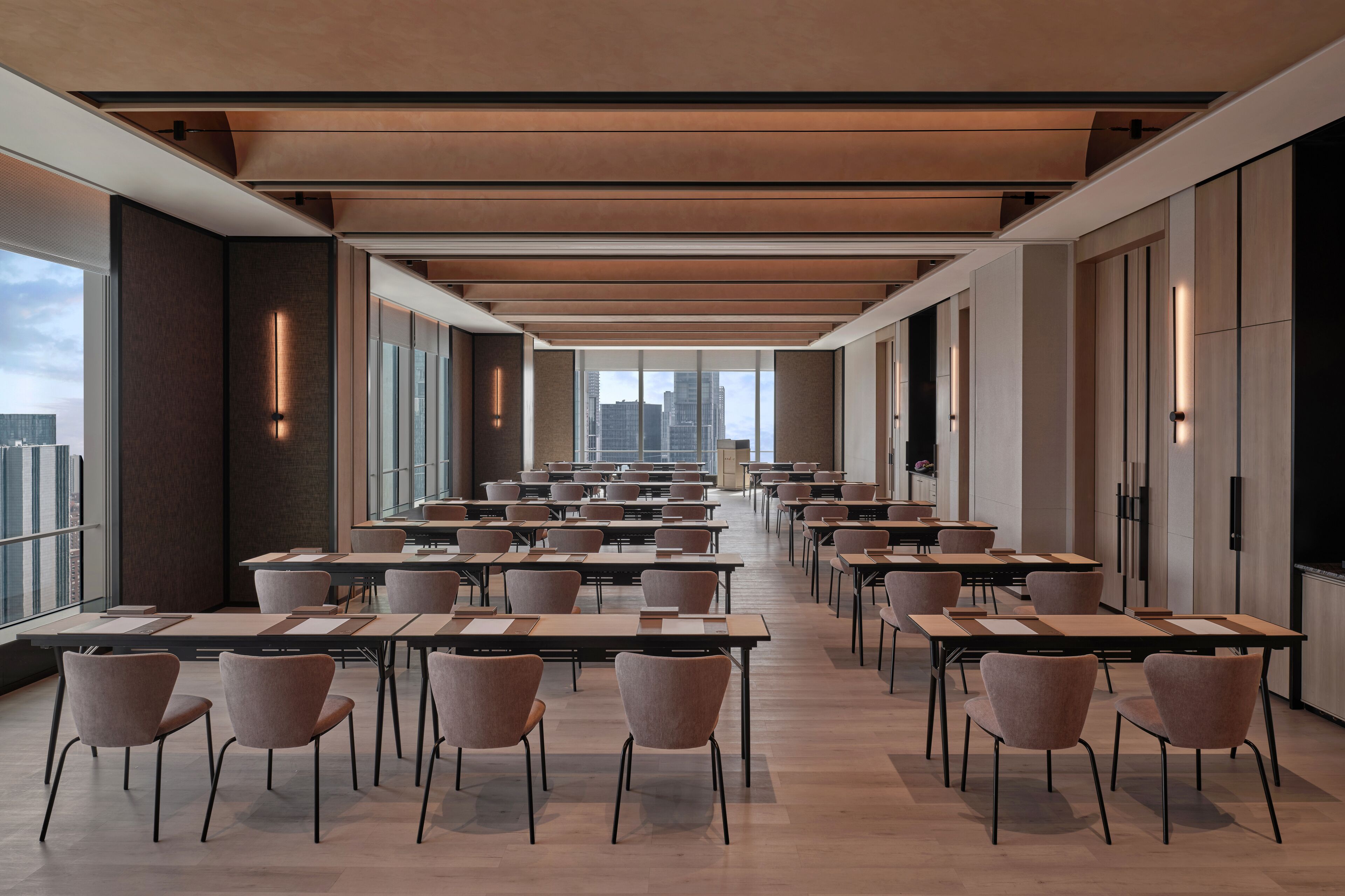Una sala de conferencias moderna con filas de mesas y sillas, ventanas amplias que ofrecen vista a la ciudad, y un diseño minimalista.