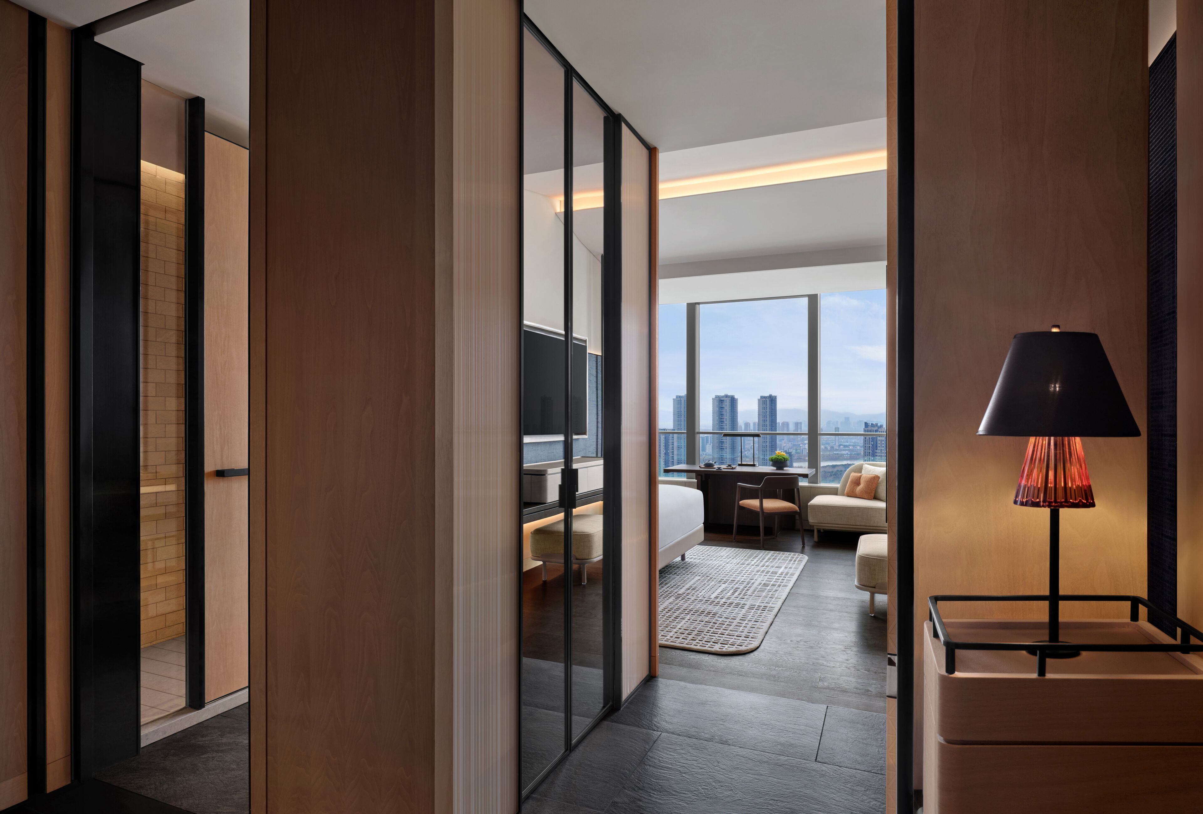 A modern hotel room with a city view, a desk, a bed, and a large window.