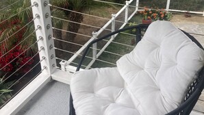 Un balcón con una silla blanca acolchada, baranda de metal y vista a una calle residencial.