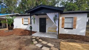 Ein kleines, eingeschossiges Haus mit schwarzem Dach und weißen Wänden, Hausnummer 2526. Es besitzt eine Veranda mit einer Bank, einem Blumentopf und einen kleinen Gartenbereich mit Mulch und Pflastersteinen.