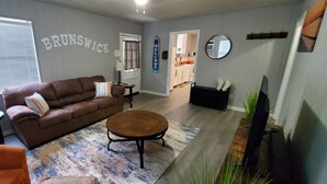 Ein Wohnzimmer mit braunem Sofa, einem runden Couchtisch und einem kleinen, orangefarbenen Sessel. Ein Fenster mit Jalousien, eine Lampe und ein wandmontierter Spiegel sind ebenfalls zu sehen.