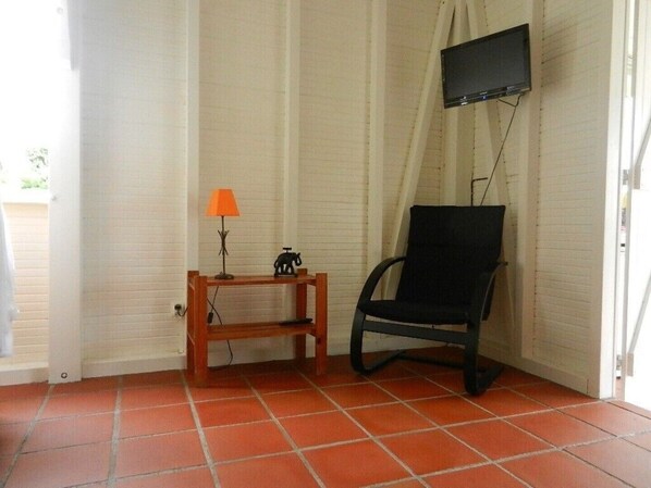 Ett rum med ett kaklat golv, en svart stol, ett litet träbord med en lampa och en liten statyett, och en tv som är monterad på väggen.