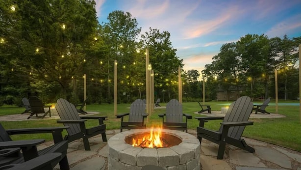 A stone fire pit surrounded by Adirondack chairs in a landscaped outdoor area with trees and string lights. - Cozy Poconos Cabin in the Pines | Firepit & Pickle (Barrett Township)