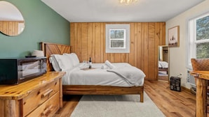 A bedroom with a wooden bed, a small bedside table with a lamp, a window with blinds, and a view of the outdoors. - Cozy Poconos Cabin in the Pines | Firepit & Pickle (Barrett Township)