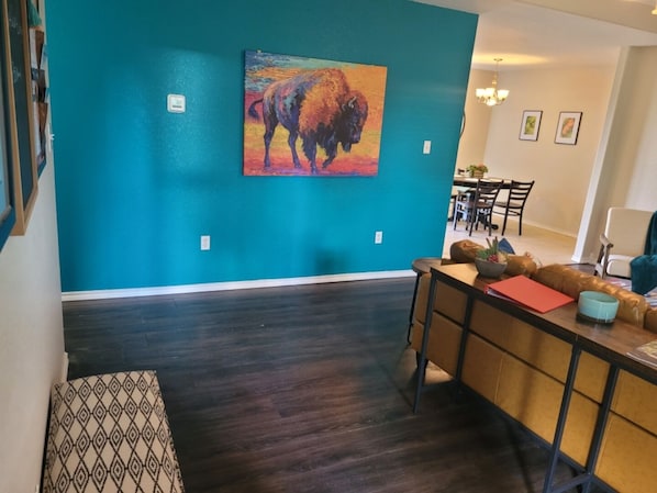 A modern living room with a teal accent wall, a dining area with a wooden table and chairs, and a large painting of a bison.