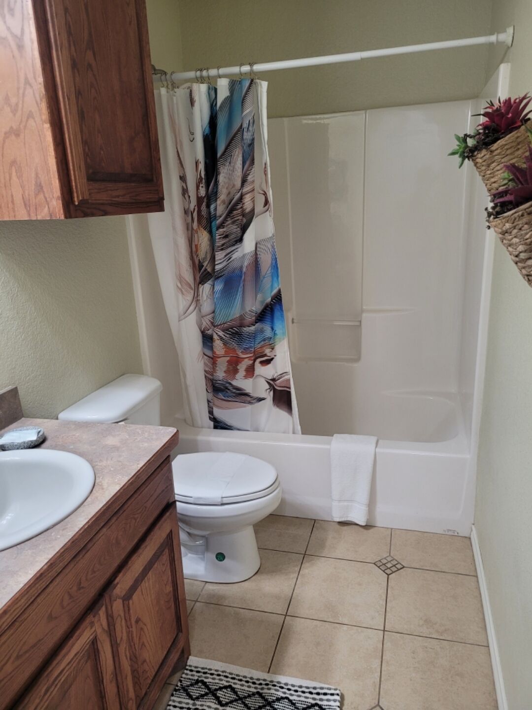 A bathroom with a wooden vanity, a white toilet, a shower curtain with a leaf pattern, and a small plant on a shelf.