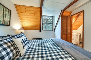 Een slaapkamer met een geruit beddegoed, houten balken in het plafond, een raam en een deur naar een badkamer.