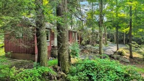 A rustic cabin surrounded by dense forest, with a stone pathway leading to the entrance. - Cozy & Chic Pocono Cabin | Firepits | Pickleball (Barrett Township)
