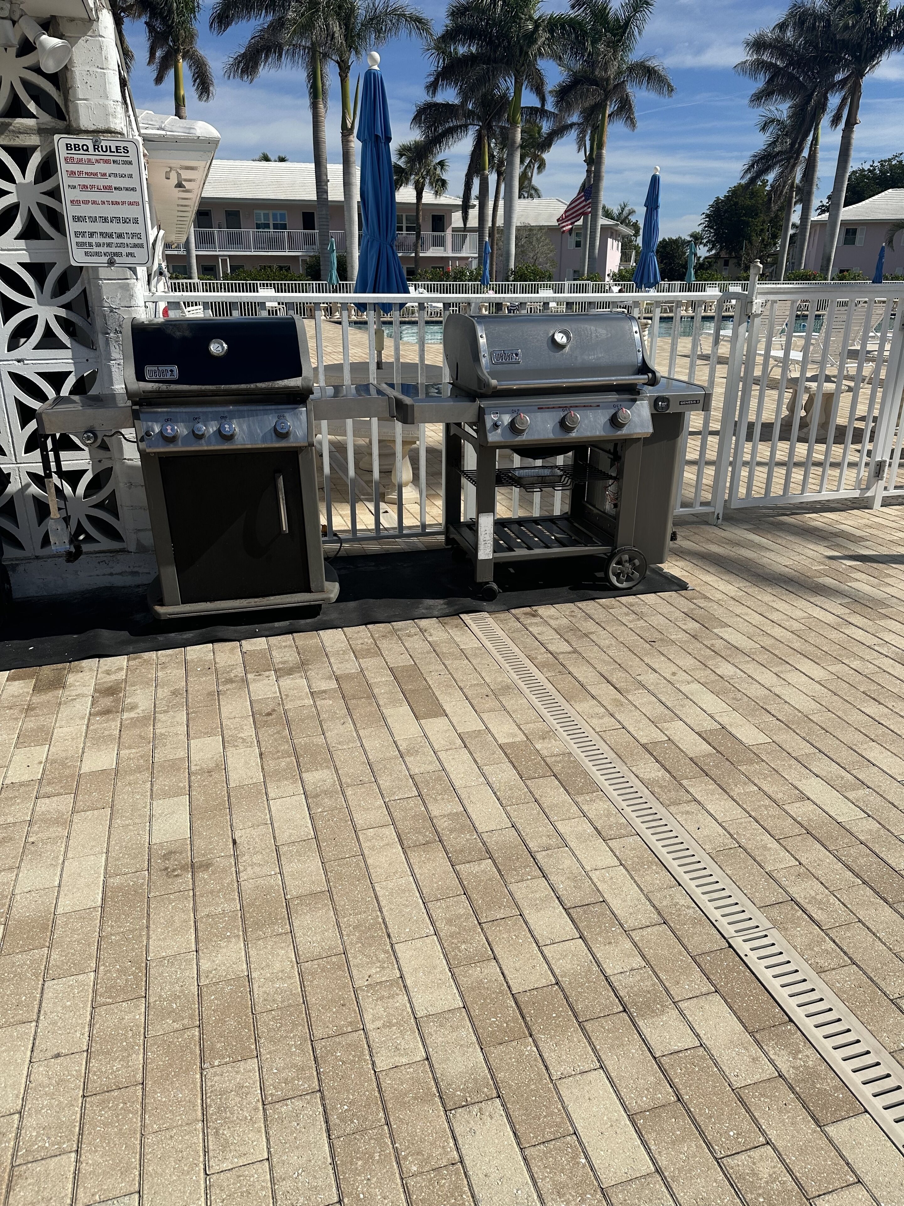 An outdoor grill area with a sign detailing BBQ rules, surrounded by a white fence and palm trees.
