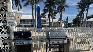 An outdoor grill area with a sign detailing BBQ rules, a brick patio, and a white fence with palm trees in the background. - Fully remodeled 1/1 apartment (Marco Island)