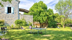 Un edificio de piedra con cancha de tenis, mobiliario exterior y un árbol grande.
