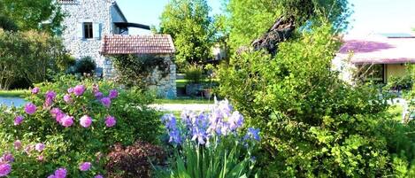 A stone house with a tiled roof, surrounded by lush greenery and flowering plants.