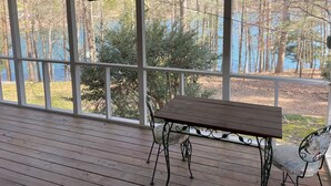 Una terraza de madera con una mesa y sillas, amplias ventanas que ofrecen vistas a árboles y un cuerpo de agua.
