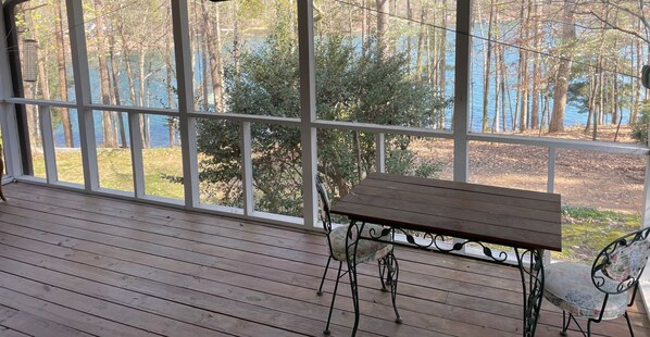 A wooden deck with a table and chairs, large windows offering a view of trees and a body of water.